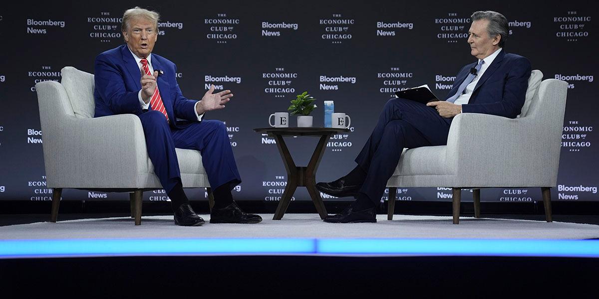 En bild på Donald Trump på scenen under en intervju med Bloomberg News. Trump och intervjuaren sitter mitt emot varandra i ljusa tygfåtöljer. Båda männen har blå kostym. Trump gestikulerar och tittar ut mot publiken.