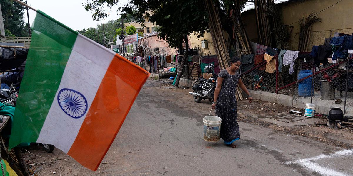 En kvinna som går i ett slumområde passerar Indiens flagga som hänger från en byggnad. Hon bär en hink med vatten.