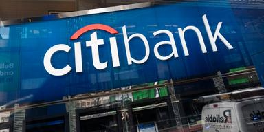 En bild som visar Citibanks logga ovanför glasentrén till deras bankkontor i New York. Texten är vit mot en blå bakgrund. Byggnader och en vit skåpbil speglas i husets glasfasad.