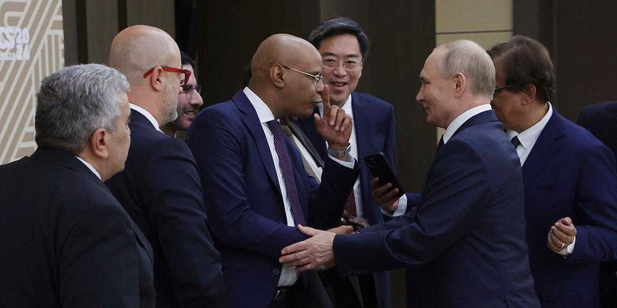 En bild som visar Rysslands president Vladimir Putin i samtal med en grupp män i blåa kostymer. Vladimir Putin, som också bär blå kostym, står i mitten och tar en av männen i hand. Mannen gestikulerar med sitt vänstra pekfinger framför sitt ansikte. Alla ser glada ut.