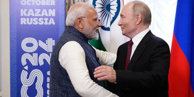 En bild som visar Indiens premiärminister Narendra Modi och Rysslands president Vladimir Putin. De står nära varandra ansikte mot ansikte och det ser det ut som att de nyss har kramat varandra.Narendra Modi bär en gråfärgad traditionell indisk väst som är hög i halsen och en vit långärmad skjorta. Vladimir Putin är klädd i svart kostym, vit skjorta och han bär en mörkröd slips. Båda männen ser nöjda ut.