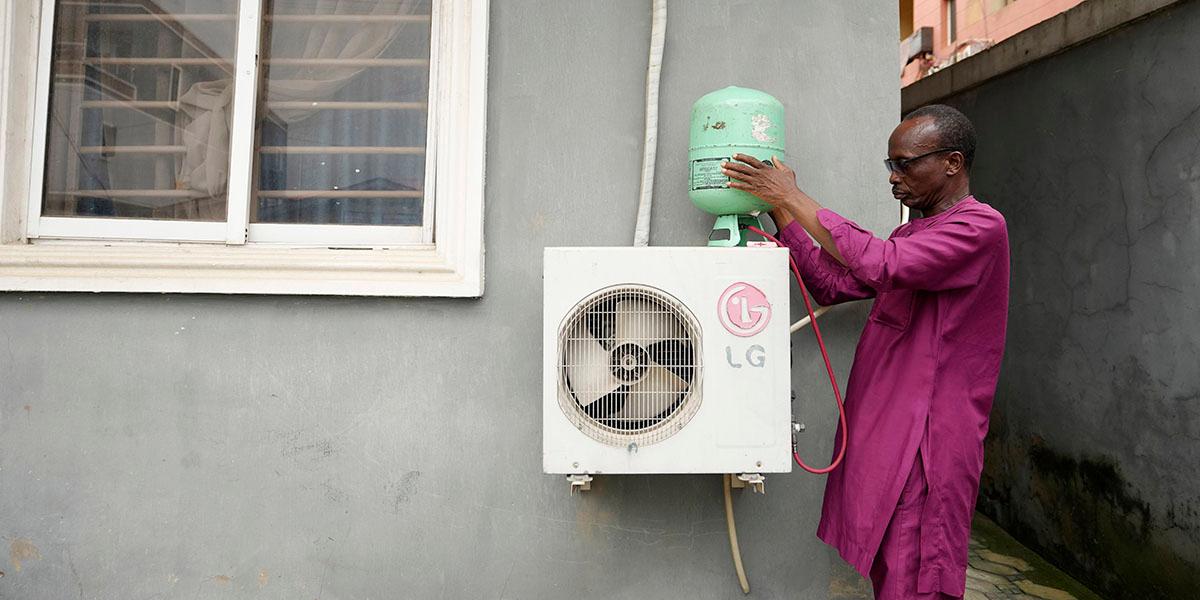 En Nigerisk man i purpurfärgade kläder gör underhåll på en klimatanläggning som är monterad nedanför ett av fönstren på ett ljusgrått hus.