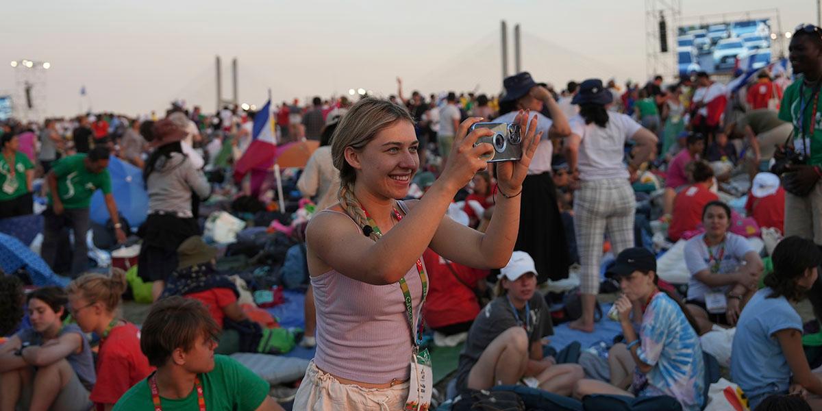 En ung kvinna står på en utomhusfestival bland många andra ungdomar och fotograferar med sin mobiltelefon.