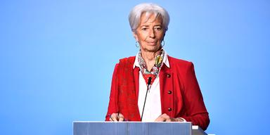 En bild på Christine Lagarde, chef för den Europeiska centralbanken. Hon står vid ett podium mot en ljusblå bakgrund. Hon har grått hår och är iklädd en röd kavaj, vit blus och en röd- och gulmönstrad scarf.