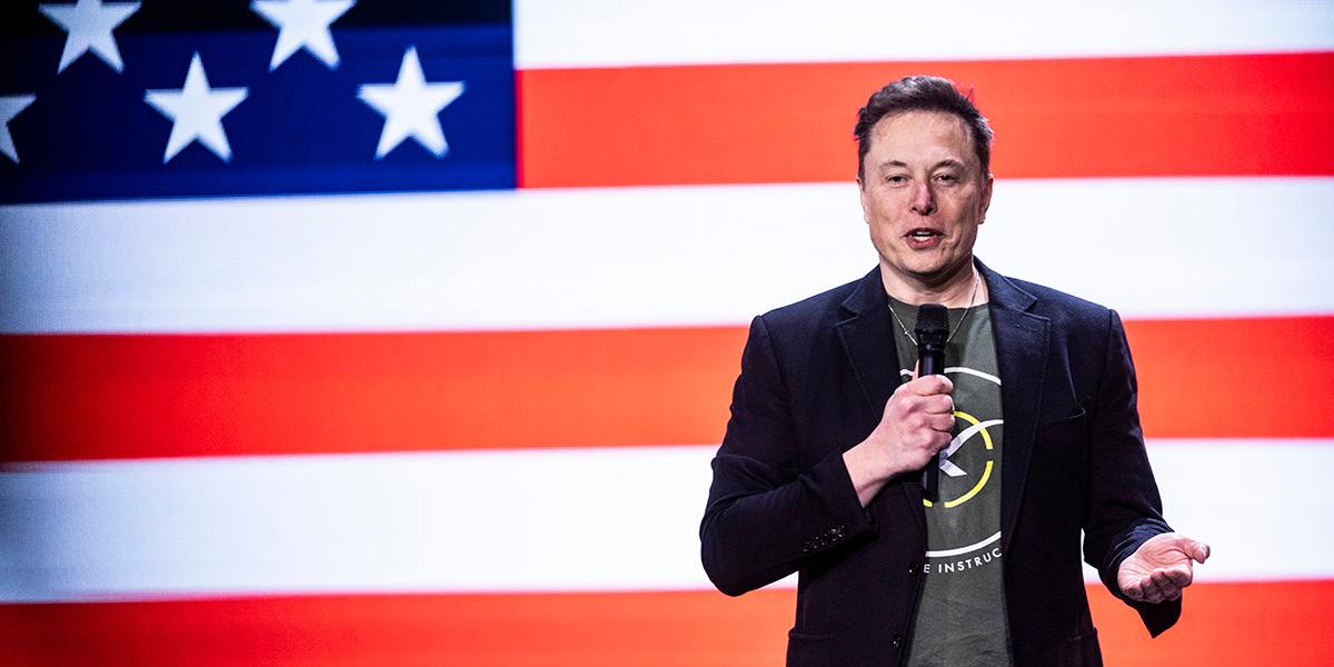 En bild på entreprenören Elon Musk i svart kavaj och en T-shirt med tryck framför en storbildsskärm som visar den amerikanska flaggan. Bilden är tagen under ett uppträdande och han håller en mikrofon i handen.