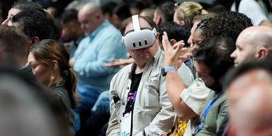 En närbild på publiken under en av Metas event där de lanserar nya produkter. Mitt i bilden sitter en medelålders man i beige rock som har på sig ett par vita VR-glasögon. Han verkar titta på en person bredvid honom som pratar och gestikulerar.