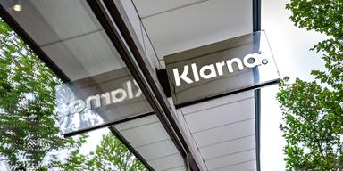 Bilden visar en spegelblank skylt med betalföretaget Klarnas logotyp i vit text. Skylten sticker ut från fasaden och den är monterad ovanför glasentrén till företagets kontor i Stockholm.