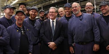 En bild på Storbritanniens premiärminister Keir Starmer under ett besök på en fabrik i Chester. Han står i mitten av bilden iklädd marinblå kostym. Runt honom står manliga arbetare iklädda mörkblåa overaller, kepsar och skyddsglasögon. Alla på bilden ser glada ut.