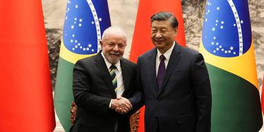 En bild på Brasiliens president Luiz Inácio Lula da Silva tillsammans med Kinas president Xi Jinping. De båda männen skakar hand framför kinesiska och brasilianska flaggor. De ser glada ut.