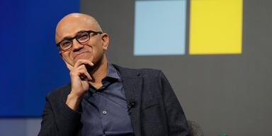 En närbild på Microsofts vd, Satya Nadella, i svart kavaj och mörkgrå skjorta. Han sitter på en scen under ett framträdande och i bakgrunden syns företagets logotyp. Han vilar ansiktet i ena handen medan han leende tittar in i kameran.