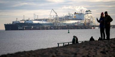 En bild på stora gas-tankers som ligger i en hamn. I förgrunden syns siluetten av två människor som tittar på fartygen.