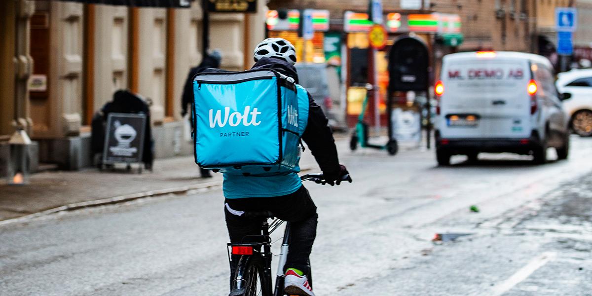 En man som arbetar för Wolt cyklar i en stadsmiljö. Han har en ryggsäck med företagets logga.